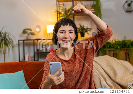 Woman browsing smartphone winning lottery celebrating success victory play game sitting on sofa 125154731