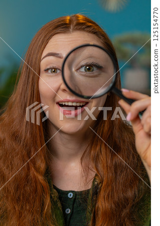 Amazed woman holds magnifying glass near face looking at camera with big zoomed eye analyzing wow 125154770