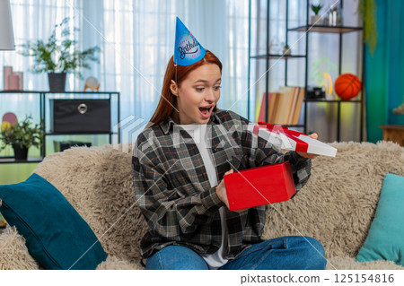 Woman wearing party hat opening birthday gift looking pleasantly surprised and emotional happy Woman wearing party hat opening birthday gift looking pleasantly surprised and emotional happy 125154816