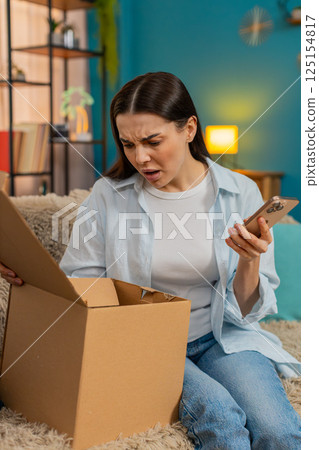 Young woman sitting on sofa unpacking delivery finding wrong item disappointed shaking head sadly Young woman sitting on sofa unpacking delivery finding wrong item disappointed shaking head sadly 125154817