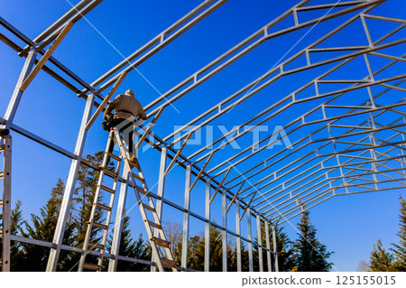 Construction frame worker climbs ladder to assemble metal framing of building under a blue sky. Construction frame worker climbs ladder to assemble metal framing of building under a blue sky. 125155015