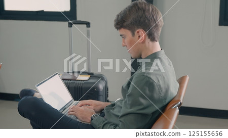 Young handsome man with a suitcase sitting working his laptop at airport terminal hall while waiting landing for her flight, tourist men in international airport, travel trip 125155656