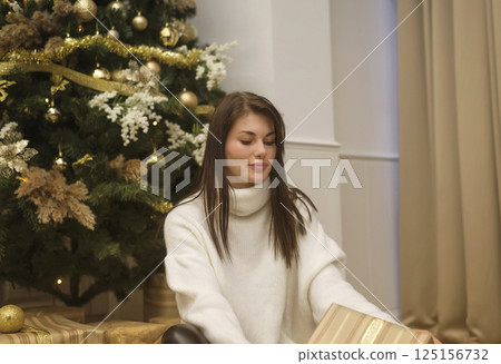 A cute young woman is sitting in front of a Christmas tree and golden boxes with gifts 125156732
