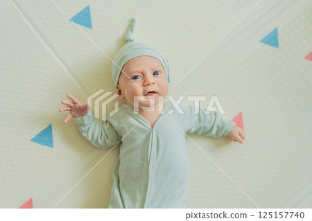 Adorable baby in a green hat lying on a play mat. Cozy newborn moment with soft textures and bright colors. Infant development, sensory play, and early childhood concept Adorable baby in a green hat lying on a play mat. Cozy newborn moment with soft textures and bright colors. Infant development, sensory play, and early childhood concept 125157740