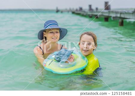 Baby floating in a neck ring in turquoise water with mom and older brother nearby. Family bonding and early swimming concept Baby floating in a neck ring in turquoise water with mom and older brother nearby. Family bonding and early swimming concept 125158481