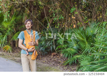 Father holding his little baby in a yellow sling in the park. Warm and loving family moment. Babywearing, fatherhood, and parent-child bonding concept Father holding his little baby in a yellow sling in the park. Warm and loving family moment. Babywearing, fatherhood, and parent-child bonding concept 125158484