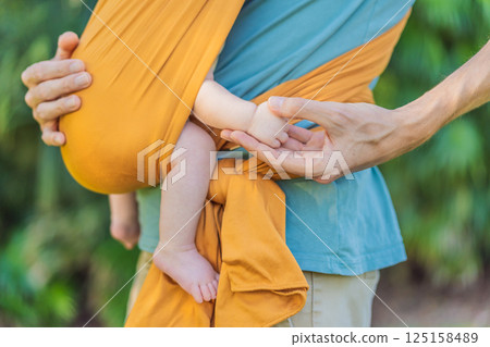 Father holding his little baby in a yellow sling in the park. Warm and loving family moment. Babywearing, fatherhood, and parent-child bonding concept Father holding his little baby in a yellow sling in the park. Warm and loving family moment. Babywearing, fatherhood, and parent-child bonding concept 125158489