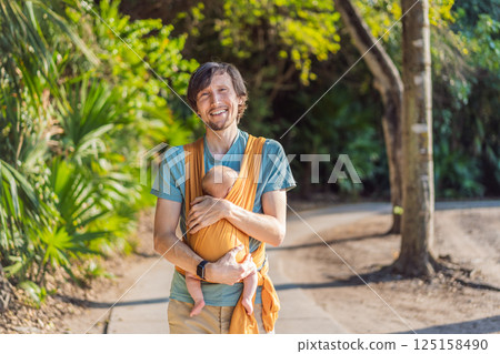 Father holding his little baby in a yellow sling in the park. Warm and loving family moment. Babywearing, fatherhood, and parent-child bonding concept Father holding his little baby in a yellow sling in the park. Warm and loving family moment. Babywearing, fatherhood, and parent-child bonding concept 125158490