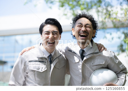 Image of a worker in construction management, etc., looking up in work clothes in front of a building under a blue sky, looking at the camera with his arm around his shoulder 125160855