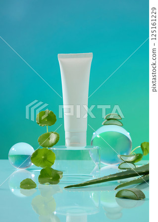 Gotu kola leaves stacked high are placed next to a glass pedestal. On top of the pedestal is an unlabeled tube of cosmetics. Aloe vera plant stems are pointed with thorns running along both sides. 125161229