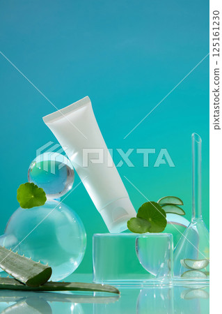 An unlabeled tube of cream stands on a square glass pedestal. The tube leans against a glass ball. The small glass ball stacked on top of the larger one, with a green pennywort leaf for decoration. 125161230