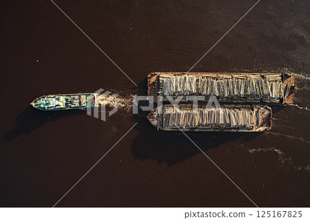 Top view: a tugboat with a barge carries timber along the river, cargo transportation by water Top view: a tugboat with a barge carries timber along the river, cargo transportation by water 125167825