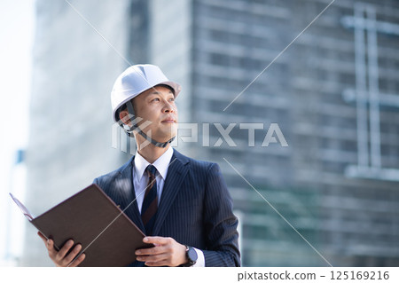 A man in a suit standing at a construction site with a document in his hand A man in a suit standing at a construction site with a document in his hand 125169216