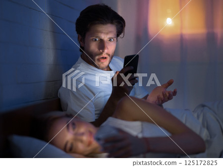 Cheating. Shocked Husband Catching Wife Chatting On Phone With Lover Lying In Bed At Night. Low Light, Selective Focus 125172825