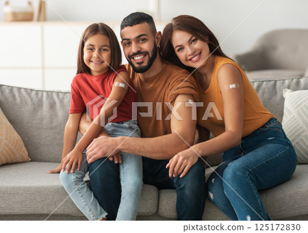 Coronavirus Vaccination Advert. Happy Vaccinated Family Of Three People Showing Arm With Sticking Plaster After Covid-19 Vaccine Vax Injection Posing Sitting On Couch In Living Room, Smiling To Camera 125172830