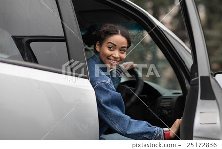 Are you going with me. Friendly smiling millennial african american female driver opens door and looks for fellow traveler, sitting in silver car or lady gets out of auto and looks back, copy space 125172836