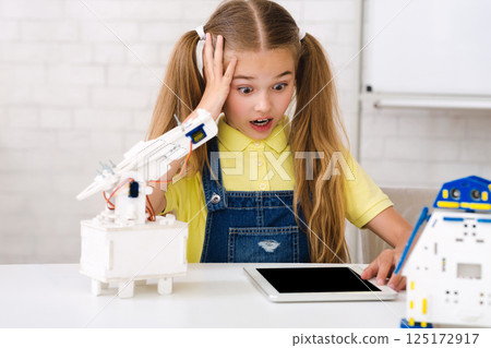 A young girl with pigtails looks surprised and concerned as she works on a robotics project. She is surrounded by robot parts and a tablet, suggesting she is following instructions 125172917