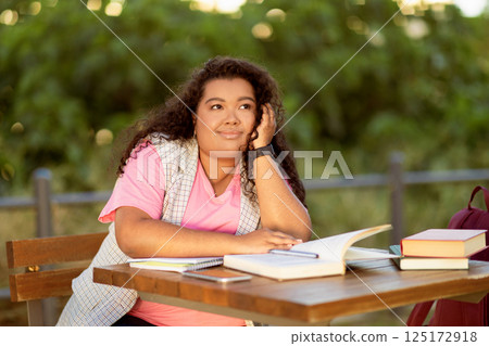 A student sits at a table surrounded by books, looking thoughtful while studying outdoors on a sunny afternoon, enjoying the greenery around her as she contemplates her work. 125172918