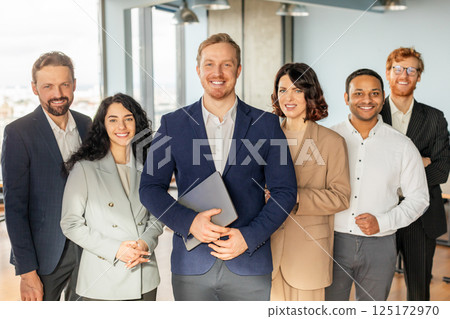 A group of smiling business professionals stand together in an office setting. The man in the center of the group is holding a laptop, while the others stand around him with their arms crossed 125172970