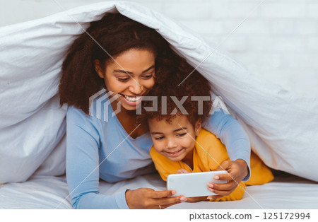 African American Mother And Daughter Using Smartphone Lying In Cozy Bed Covered With Blanket Relaxing At Home. Selective Focus African American Mother And Daughter Using Smartphone Lying In Cozy Bed Covered With Blanket Relaxing At Home. Selective Focus 125172994