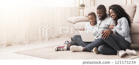 Planning vacation together. African father, mother and daughter using laptop at home, sitting on floor, copy space 125173239