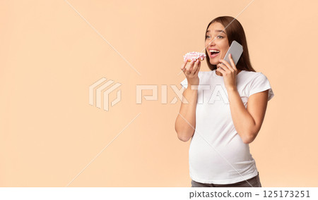 Excited Pregnant Woman Chatting On Cellphone Eating Doughnut Standing On White Studio Background. Copy Space 125173251