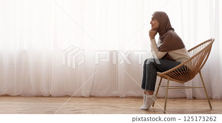 Morning Harmony. Muslim girl in hijab relaxing in wicker chair at home, resting head on hands and daydreaming against window, side view 125173262