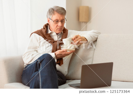 Senior Man Using Laptop Computer Having Coffee Sitting On Sofa At Home On Weekend. Selective Focus Senior Man Using Laptop Computer Having Coffee Sitting On Sofa At Home On Weekend. Selective Focus 125173364
