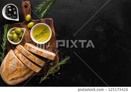 Olive oil and olives in bowls, sliced bread and rosemary on cutting board over concrete background, top view, copy space 125173428
