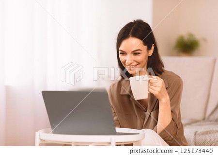 Good Morning. Smiling student drinking coffee at home and using laptop, watching video, chatting with friends, copyspace 125173440