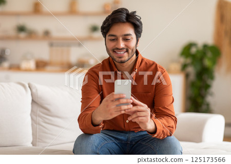 Arab man in an orange shirt and blue jeans sits on a white couch while looking at a smartphone. He is smiling and appears to be happy or engaged with what he is viewing on his phone. 125173566