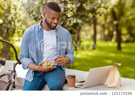 Lunch Outdoors. Smiling Black Guy Eating Vegetable Salad And Drinking Coffee While Relaxing With Laptop In Park During Break At Work 125173599