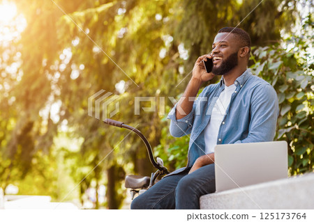 Cell Communication. Portrait Of Happy Handsome Afro Man Talking On Cellphone While Sitting With Laptop And Bicycle In Park Cell Communication. Portrait Of Happy Handsome Afro Man Talking On Cellphone While Sitting With Laptop And Bicycle In Park 125173764