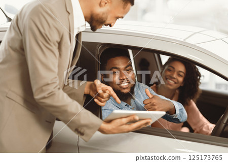 Buying New Car. Happy Black Couple Gesturing Thumbs Up Talking With Seller Approving New Automobile In Dealership Store 125173765