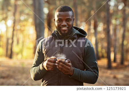 Positive african man drinking tea at forest, camping and hiking concept, empty space 125173921