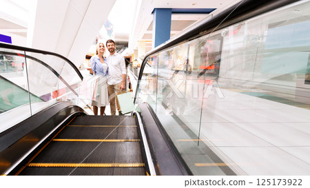 A couple stands on an escalator in a modern shopping mall, both smiling and holding shopping bags. The escalator is moving upward, with a clear glass side panel and a black metal handrail. 125173922