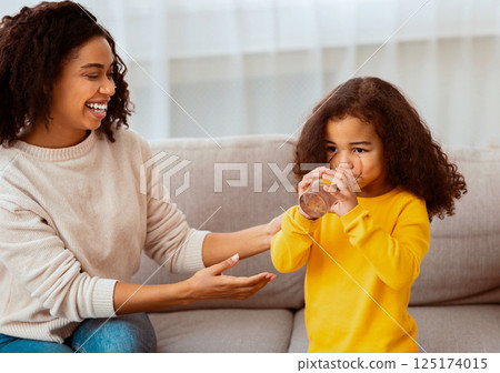 Mother's Care. Joyful African American Woman Feeding Little Daughter Giving Her Glass Of Water Sitting On Sofa At Home. 125174015