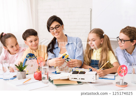 A female teacher, wearing glasses, guides four young students in a classroom setting, teaching them about robotics. The students are all engaged in the lesson 125174025
