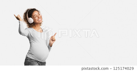 Joyful afro pregnant woman listening music in headphones and dancing over white studio background 125174029