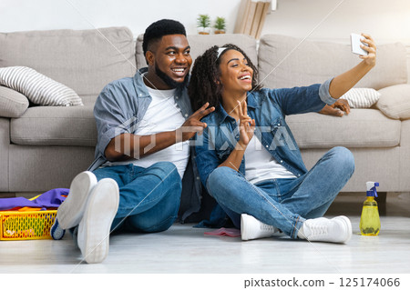 Positive black couple taking selfie on smartphone after tidying apartment together, sitting on floor, resting after spring cleaning, free space Positive black couple taking selfie on smartphone after tidying apartment together, sitting on floor, resting after spring cleaning, free space 125174066