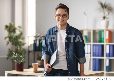 Portrait Of Handsome Young Male Entrepreneur Holding Laptop And Posing In Office Interior, Confident Millennial Man In Casual Clothes Standing Near Desk And Smiling At Camera, Copy Space 125174100