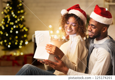 Happy afro couple opening Christmas gift box in living room, in front of xmas tree 125174201