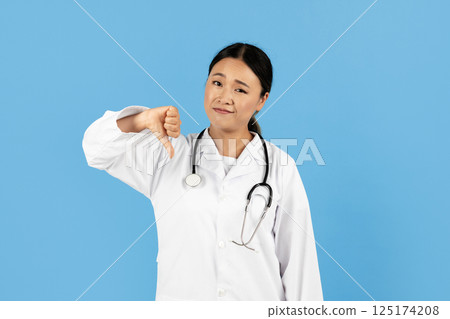 A healthcare professional Asian lady in a white coat expresses disapproval with a thumbs down gesture. She has a stethoscope around her neck, set against a bright blue background. A healthcare professional Asian lady in a white coat expresses disapproval with a thumbs down gesture. She has a stethoscope around her neck, set against a bright blue background. 125174208
