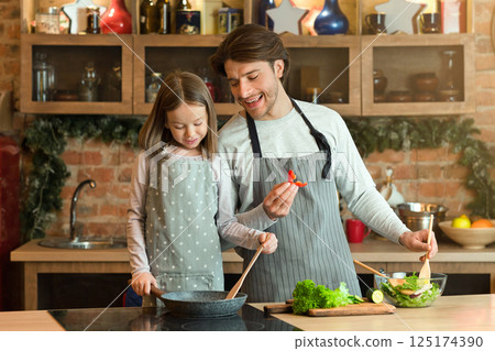 Young man and his little daughter cooking together in kitchen, preparing healthy meals with frying pan, enjoying homemade food, making vegetable salad for lunch, having fun at home, free space 125174390