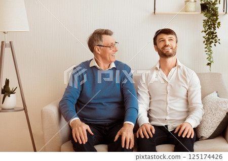 Cheerful Adult Son And Senior Father Talking And Smiling Sitting On Sofa At Home. Relationship With Elderly Parents. Selective Focus 125174426