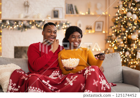 Free time, favorite movie and self-isolation at home. Millennial funny african american husband and wife with remote control and popcorn covered with blanket and watching tv in interior with tree Free time, favorite movie and self-isolation at home. Millennial funny african american husband and wife with remote control and popcorn covered with blanket and watching tv in interior with tree 125174452