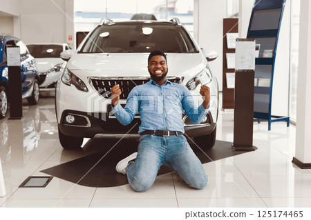 Buying New Car. Excited Black Guy Shaking Fists Standing On Knees Near Auto In Dealership Store Buying New Car. Excited Black Guy Shaking Fists Standing On Knees Near Auto In Dealership Store 125174465