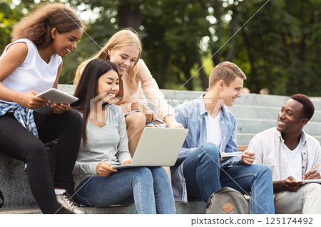 Multiethnic group of positive students sitting on stairs outdoors and doing project together Multiethnic group of positive students sitting on stairs outdoors and doing project together 125174492