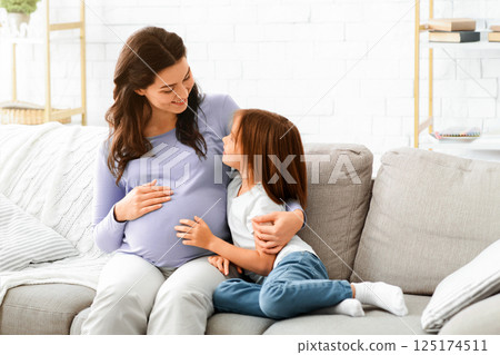 Pregnant woman and little daughter sitting on sofa at living room and having conversation, free space Pregnant woman and little daughter sitting on sofa at living room and having conversation, free space 125174511