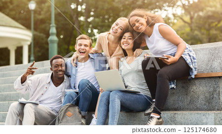 Happy friends international students posing while studying with laptop in city, sitting on stairs, empty space 125174680